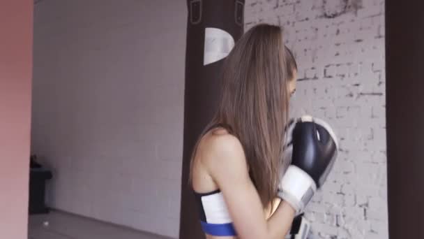 La chica se está preparando para una competición de boxeo y entrena golpes en un saco de boxeo en un amplio gimnasio — Vídeos de Stock