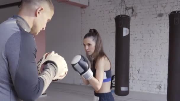 Średnie ujęcie młodej kobiety ćwiczącej boks na siłowni i uderzającej swojego instruktora, która unika ciosów i kontroluje proces treningowy — Wideo stockowe