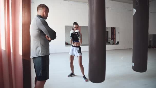 Egy szigorú edző nézi, ahogy a női kickboxoló diákja bokszzsákon gyakorolja az ütést egy tágas edzőteremben. — Stock videók