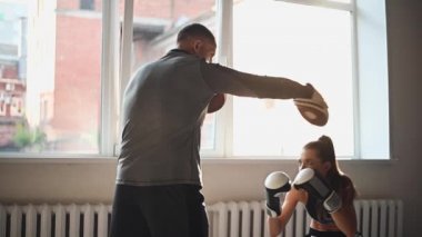 Ağır çekimde genç bir kadın spor salonunda boks antrenmanı yapıyor ve eğitmenini yumrukluyor, o da yumruklarından kaçıyor ve antrenman sürecini kontrol ediyor.