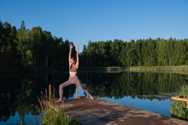 Genç kadın göldeki ahşap ranzada yoga yapıyor. Güneşli havalarda tek spor doğa üzerine sağlıklı eğitim