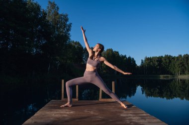 Güneşli bir günde, bir orman gölünde ruhani yoga pozu veren güzel genç bir kadın.