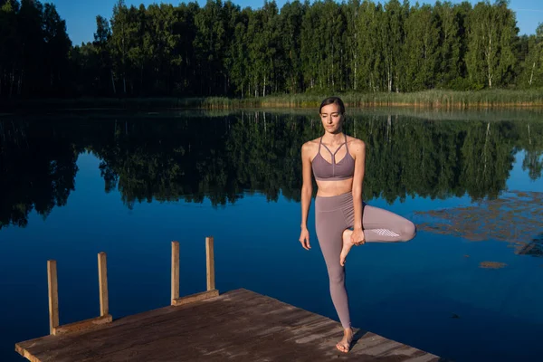 Girl doing yoga tree pose in nature. Girl yoga pose. Sports outdoors stretching on a forest lake. — Stock Photo, Image