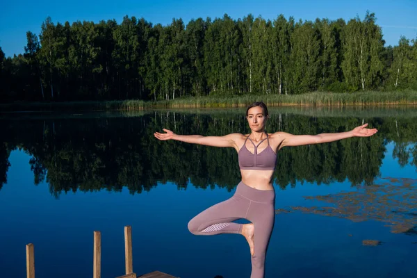 Young woman by the lake practicing yoga moves on wooden platform. Pretty young woman exercising in nature, healthy lifestyle young people positive vibes. — Stock Photo, Image
