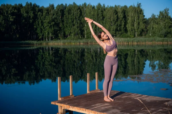 Mujer haciendo yoga en un lago del bosque. Hermosa chica haciendo ejercicios de estiramiento y flexibilidad al aire libre — Foto de Stock