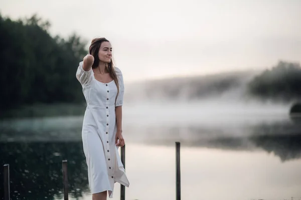 Tender romantiske sentimental kvindelig dame om morgenen på en træ mole nær den tågede flod i en hvid kjole - Stock-foto