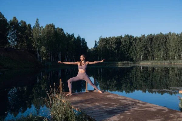 Young woman on wooden pier above forest lake scenery, folds her arms in a namaste gesture. Woman arms outstretched in nature. — Stock Photo, Image