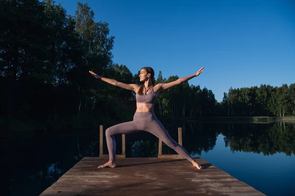 Young woman on wooden pier above forest lake scenery, folds her arms in a namaste gesture. Woman arms outstretched in nature. — Stock Photo, Image