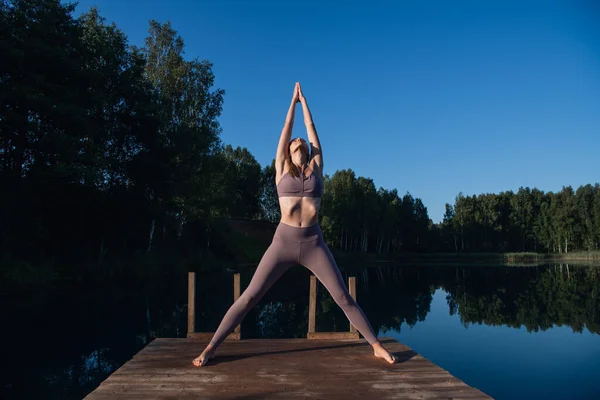 Eine junge Frau am See, die Yoga praktiziert, bewegt sich auf einer Holzplattform. Hübsche junge Frau, die in der Natur Sport treibt, gesunder Lebensstil, positive Stimmung bei jungen Menschen. — Stockfoto