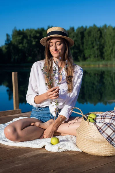 Portrait beautiful stylish romantic retro girl holding flower branch in hand while sitting at pier near the lake outdoors in nature. Stock Picture