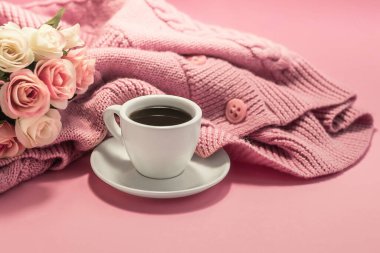 Morning coffee. A cup of coffee with a saucer and a warm sweater on the background of a bouquet of roses flowers on a pink background. Cozy morning. Copy space.