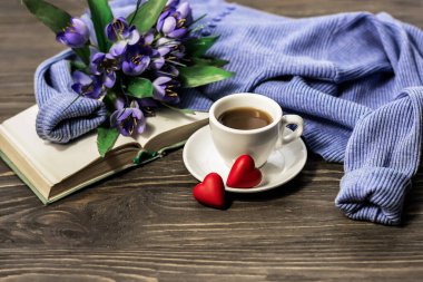 Morning coffee. A cup of coffee with a saucer and two sweet hearts on a wooden table, an open book and a warm sweater on the background of a bouquet of spring flowers. Cozy morning.
