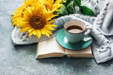 Morning coffee. Cozy composition: a cup of coffee on an open book, a blue sweater and sunflower flowers on a concrete background. Still life concept. Copy space