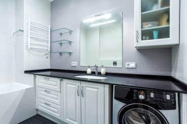 Leğen dolabı, aynası, tuvaleti, çamaşır makinesi ve taş tezgahı olan yüksek parlak bir banyo.