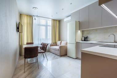 Masası, yumuşak koltukları, kanepesi ve bej bir buzdolabı olan modern bej ve gri stil bir mutfak. Döşeme kiremitleri ve kaplama. Ev aletlerinde yapılmış.