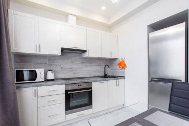 Pencerenin yanında bir sürü beyaz ve bej dolap, tezgah, lavabo, fayanslı mermer duvarın yanında mutfak aletleri olan yemek pişirme alanı..