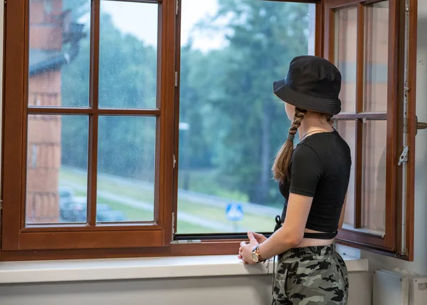 The girl is standing near the open window. Girl looking at the rain through an open window