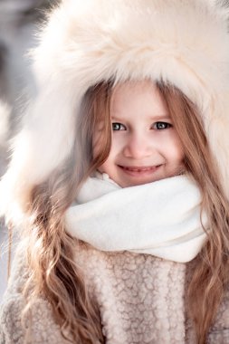 Mutlu, pozitif, neşeli, küçük bir kızın, tatlı bir çocuğun portresi. Güzel çocuk kış parkında kışın buz gibi soğuk kış gününde kürk şapkayla yürüyor. Aile tatili, moda konsepti..