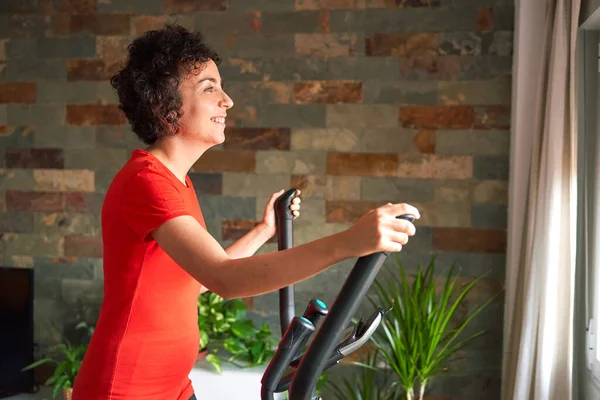 Spor salonundaki kadın spor eğitmeni, gün ışığında evde eliptik kros eğitmeni eğitimi alıyor.
