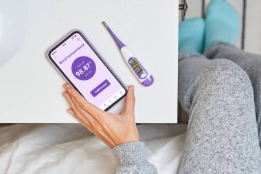 A woman records her basal temperature in the application of her smart phone thanks to the measurements of her basal thermometer to know when are the optimal days for pregnancy clipart