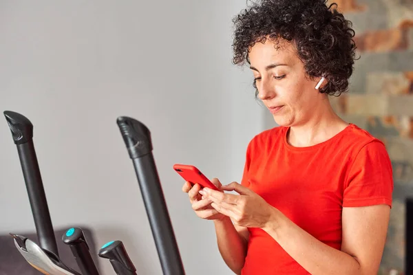 Woman exercising at home on cross training elliptical using mobile phone — Stock Photo, Image