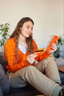 Kanepeden akıllı telefonuyla turuncu renk giyinmiş bir kadın alışveriş yapıyor.