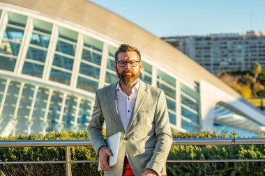 Takım elbiseli genç bir iş adamı dizüstü bilgisayarıyla arka planda modern bir bina inşa ediyor.