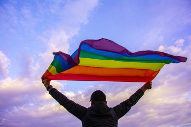 Mavi gökyüzünde Gay Gökkuşağı Bayrağı taşıyan bir adam. Biseksüel, gey, lezbiyen, transseksüel sembol.
