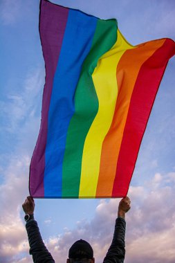 Mavi gökyüzünde Gay Gökkuşağı Bayrağı taşıyan bir adam. Biseksüel, gey, lezbiyen, transseksüel sembol.