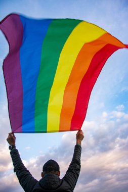 Man holding a Gay Rainbow Flag over blue summer sky. Bisexual, gay, lesbian, transsexual symbol.