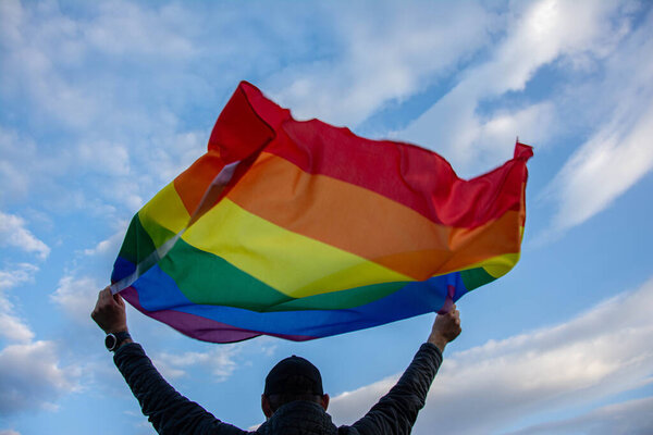 Man holding a Gay Rainbow Flag over blue summer sky. Bisexual, gay, lesbian, transsexual symbol.