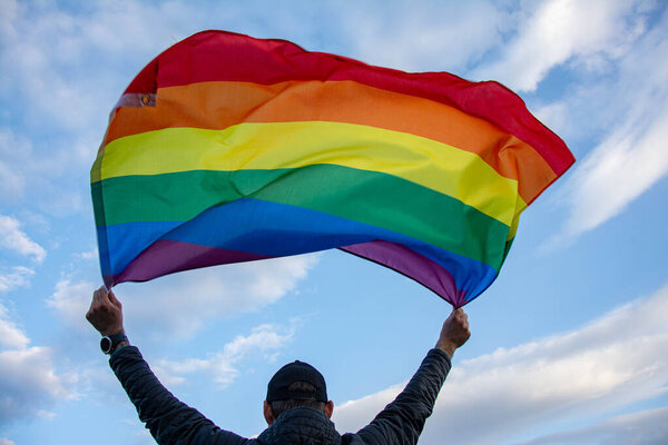 Man holding a Gay Rainbow Flag over blue summer sky. Bisexual, gay, lesbian, transsexual symbol.