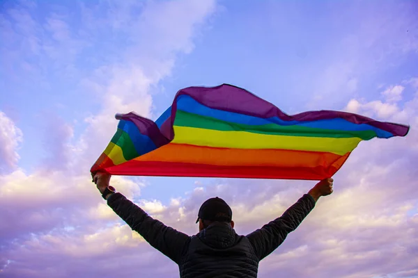 Man holding a Gay Rainbow Flag over blue summer sky. Bisexual, gay, lesbian, transsexual symbol.