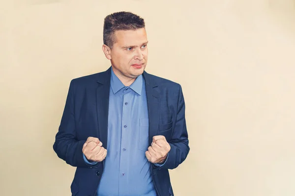 A young businessman in a blue suit clenched his hands into fists in an angry state looks to the side on a yellow background. Frustration at work. Angry emotion. copy space