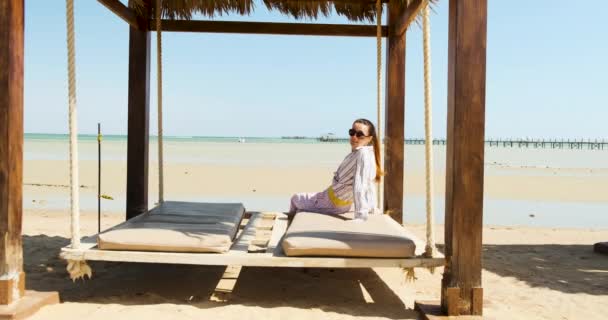 Kaukaska piękna dziewczyna w okularach przeciwsłonecznych, koszuli i spodniach siedząca na leżaku w baldachimie na plaży, ciesząca się resztą, morzem, słońcem i piaskiem. — Wideo stockowe