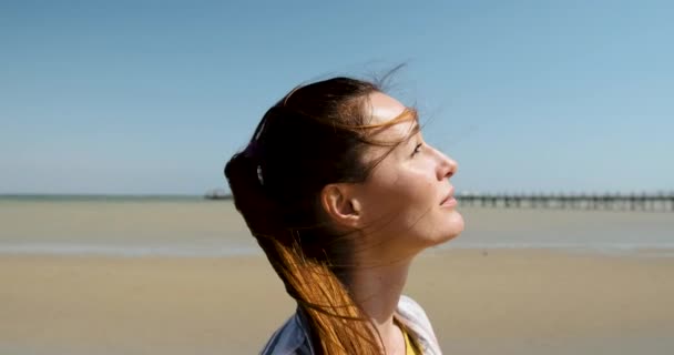 빨간 머리의 여자가 찡그리며 밝은 태양을 바라봅니다. 청명 한 날, 바다의 모래바람을 배경으로 — 비디오