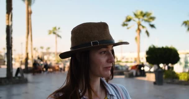 Dziewczyna w kapeluszu w cieniu, w przejrzysty dzień na tle palm, wygląda troskliwie z boku, wiatr rozwija długie włosy. Zwolnij, zbliżenie — Wideo stockowe