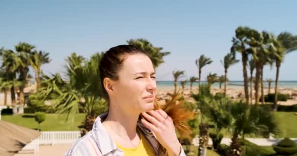 Девушка щурится от солнца, затем надевает солнцезащитные очки и наслаждается летним отдыхом в ясный день на курорте среди пальм — стоковое видео