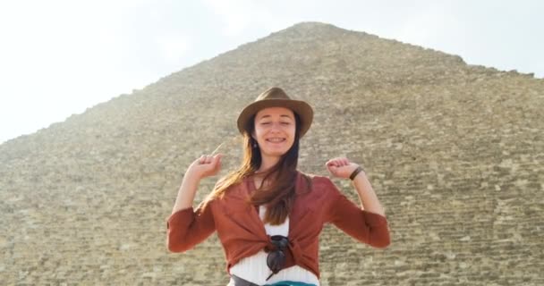 Kaukázusi lány kalapban, a nagy egyiptomi piramis előtt áll, mosolyog és boldogan integet a kezével. Közepes lövés, lassított felvétel, tiszta napos nap — Stock videók
