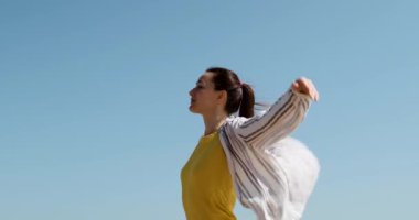 Bir kadın sarı tişört ve tişörtlü kuş gibi kollarını ve dalgalarını açık mavi gökyüzüne doğru uzatıyor. Orta boy görüntüler, parlak güneş ve güçlü rüzgar.