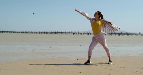Een langharig meisje staat in de wind op een zandstrand bij eb in een hand in de voorste houding. Brede beelden, schaduw op het zand, door de wind geblazen haar en een shirt — Stockvideo