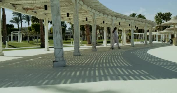 Chica camina entre las columnas, un dosel blanco de verano hecho de madera. La sombra del sol cae maravillosamente sobre el telón de fondo de las palmeras del complejo. — Vídeos de Stock