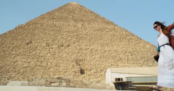 Gadis Kaukasia berambut panjang berkacamata hitam dan gaun putih berjalan melewati Piramida Agung Giza di Kairo Mesir. Tangan terpisah, hari yang cerah, tembakan lebar, gerakan lambat — Stok Video