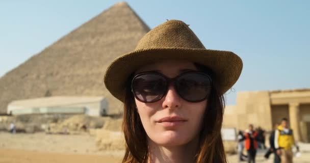 Кавказская девушка в брекетах, в шляпе в солнечных очках, улыбается на фоне Великой Пирамиды Гизы в Египте. Чистый день, крупный план, туристы на заднем плане — стоковое видео