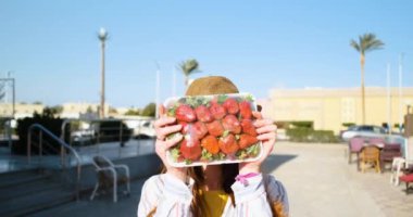 Şapkalı genç beyaz bir kız, taze çilek paketinde geziyor ve seviniyor. Yakın çekim, açık gün, yaz, neşe, ağır çekim