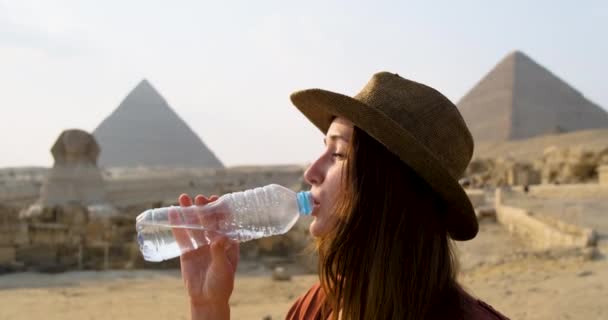 Girl in a hat drinks water from a plastic bottle with pleasure, against the backdrop of the Sphinx and the Egyptian pyramids. Clear sunny day, heat, slow motion, close up — Stock Video