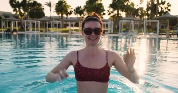 Flicka i bikini och solglasögon som hoppar i poolen. Glad, leende mot bakgrund av palmer och nedgående sol. — Stockvideo