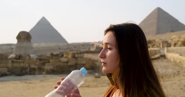 Mladá běloška s dlouhými vlasy, krásně a s potěšením, pije vodu v poušti Sahara, na pozadí sfingy a egyptských pyramid. Jasný slunečný den, vedro, zpomalený pohyb — Stock video