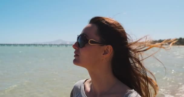 Retrato de una joven pelirroja caucásica que mira al mar con gafas de sol, el viento desarrolla el pelo largo, sonríe y disfruta de la vista del océano. Día soleado claro, cámara lenta — Vídeo de stock