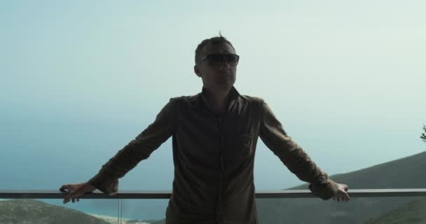Ein junger Mann, der mit Sonnenbrille im Schatten steht, hält seine Hände am Geländer des Glasgeländers und blickt langsam in die Sonne. Zeitlupe, blauer Himmel, schöne Aussicht im Hintergrund — Stockvideo